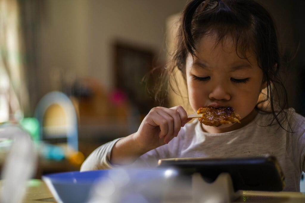 Happy asian little girl eating and watching video from smartphone.
